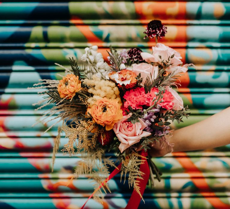 Bride holds colourful mixed bridal bouquet with pink roses tied with red ribbon infront of colourful background in streets of Liverpool