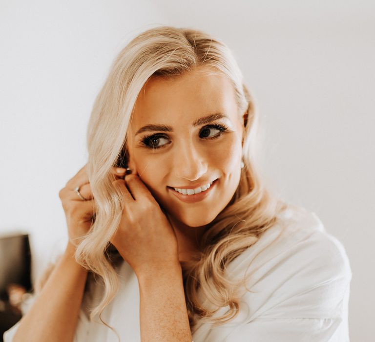 Bride with blonde curled hair and white bridal robe smiles as she puts in earrings before wedding