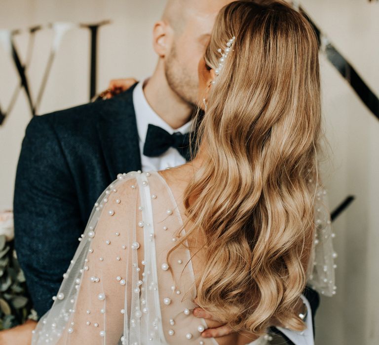 Bride with long curled hair and pearl hair clip wearing wedding dress with mesh pearl overlay kisses groom in navy tweed suit and bow tie