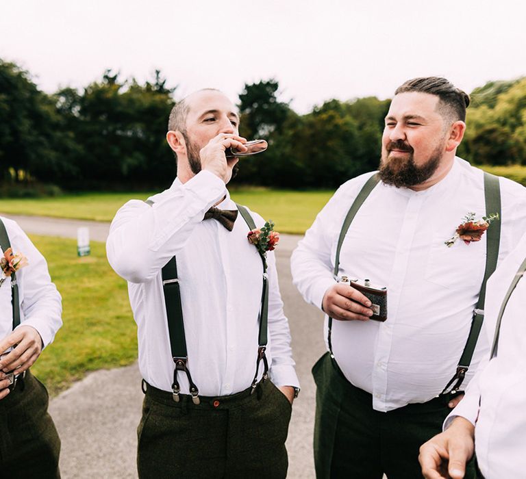 Groom with the groomsmen in matching olive green trousers and braces drinking from hip flasks 