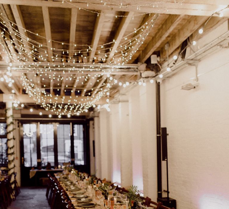 Light wood banquet tables with sage green napkins and plates with dark wooden chairs and overhead fairy lights