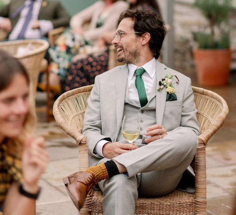 Groom sits on wicker chair in bespoke grey suit and funky socks as he socialises with wedding guests