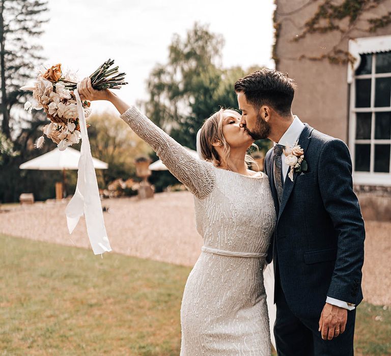 Bride in beaded dress and silver wedding shoes holds neutral wedding bouquet and kisses groom in blue suit with paisely tie