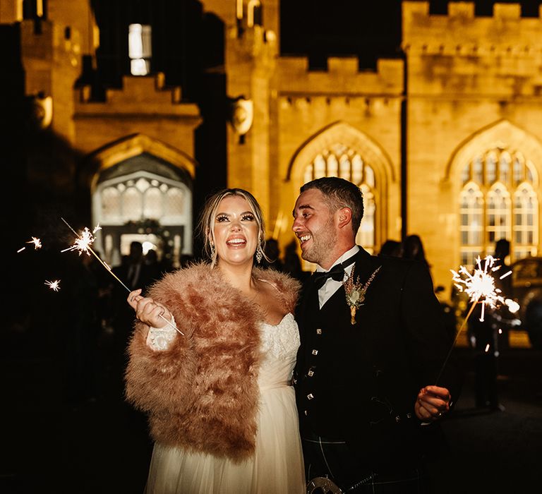 Bride looks up at fireworks and groom looks at her as they wave their own sparklers around 