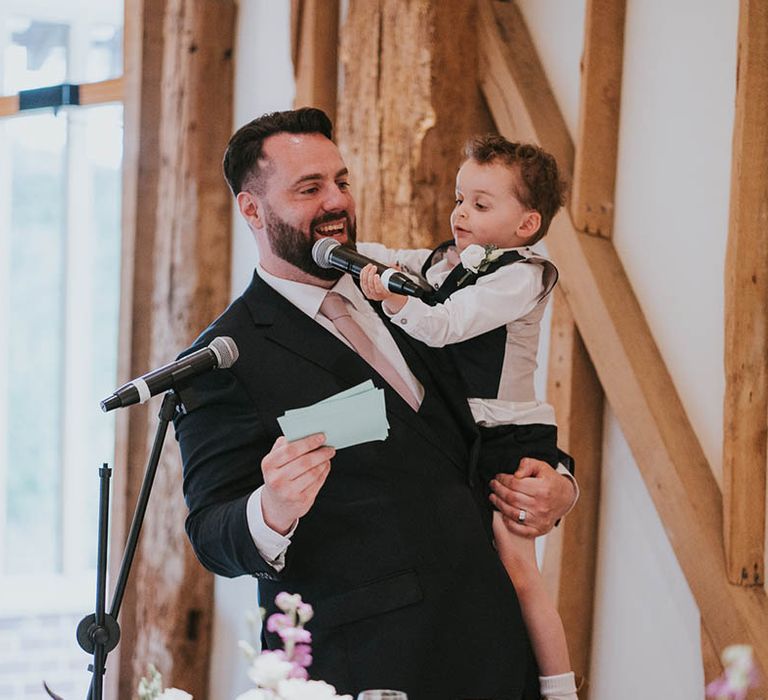 Bearded groom reads out his wedding speech as he holds onto young page boy in dark grey waistcoat 