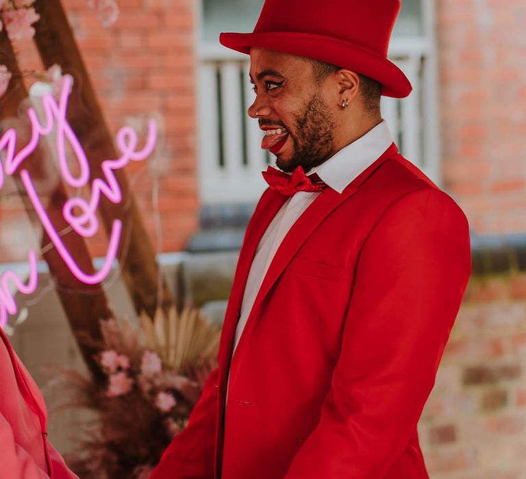 Stylish groom in a red suit and top hat wearing winged eyeliner and red lipstick makeup