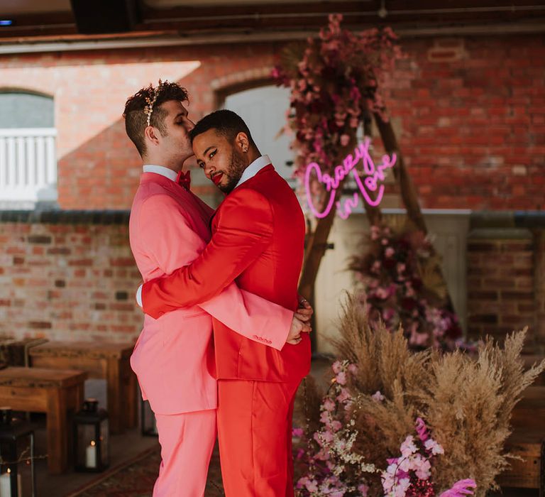 Groom in a pink suit, white boots and gold crown kissing his partners head in a red suit at Prestwold Hall Barns 