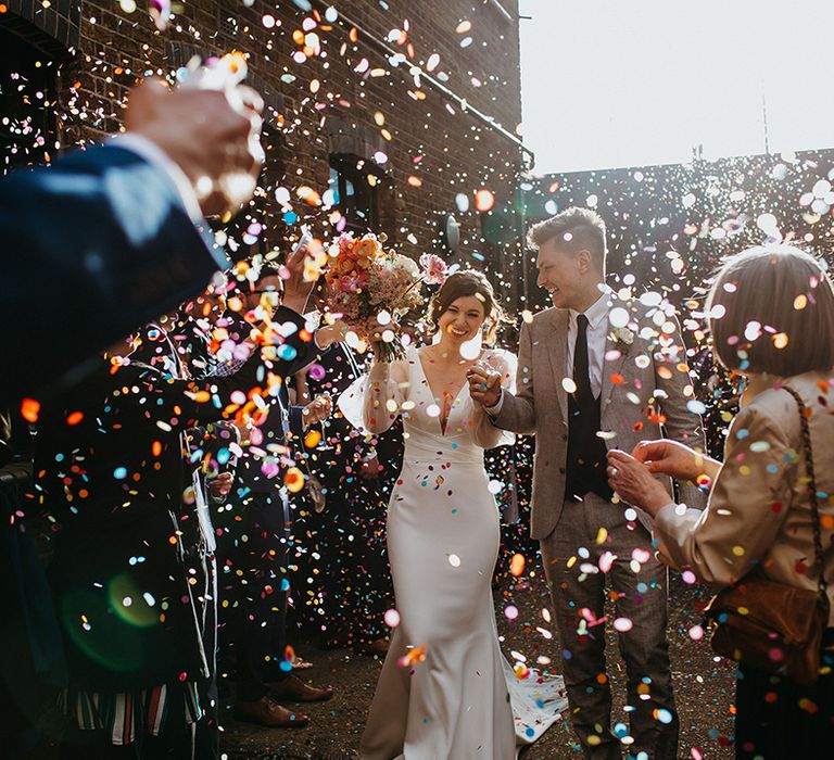 Bride and groom exit their ceremony at Loft Studios to colourful confetti with bride holding bright bouquet with giant poppy 