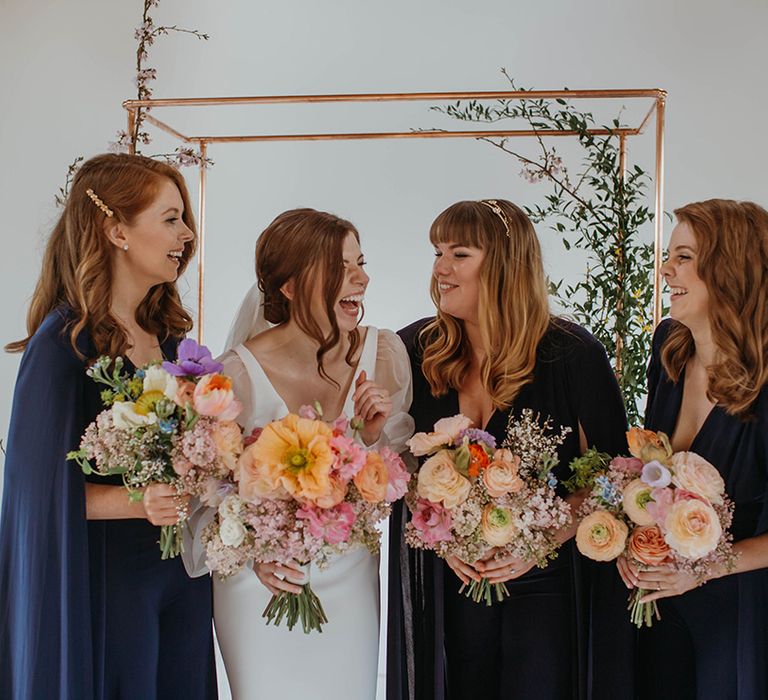 Bride and bridesmaids laugh together as they all hold colourful bright spring bouquets with giant poppy