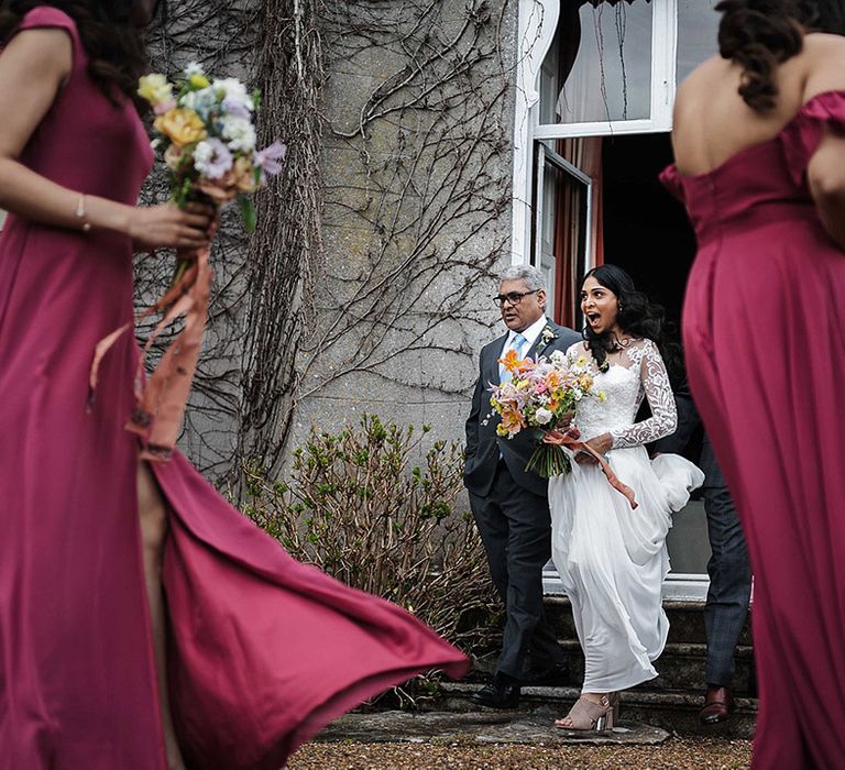 Bride walks out of the house in her lace long sleeve wedding dress and man in grey suit and baby blue tie with bridesmaids in wine colour dresses 