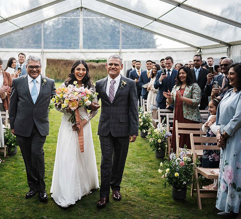 Bride in white wedding dress with long sleeves holding colourful spring bouquet is walked down the aisle by two men in grey suits