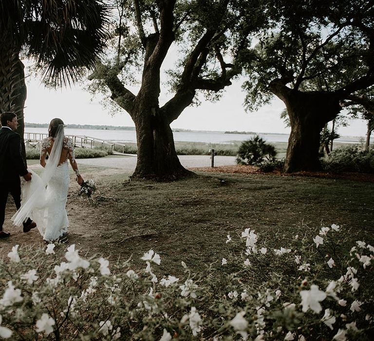 Groom walks with the bride holding her dress up as bride wears long floral illusion lace long sleeve wedding dress 