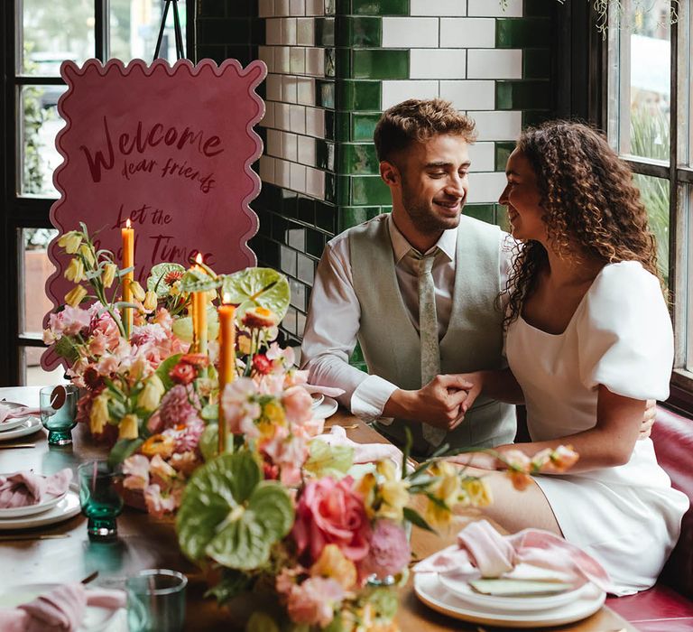 Bride in a short wedding dress and groom in a pale green suit sitting at their zesty reception at Singer Tavern with pink wedding sign and flower centrepieces 