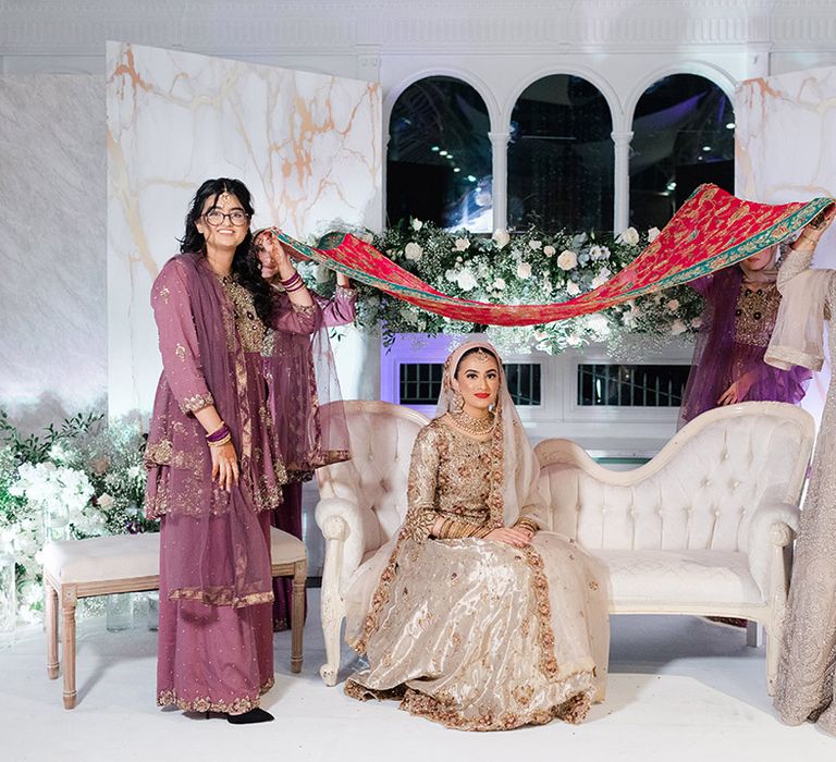 Bride sits on white sofa as wedding guests hold brightly coloured fabric above her head after Walima & Nikkah 