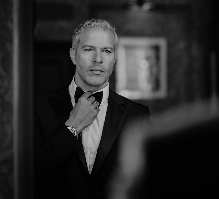 Groom in black tie adjusts his bow tie as he looks in the mirror 