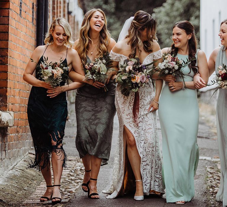 Bride in a boho lace wedding dress with bridesmaids in different shades of green dresses with small pink, red and purple flower bouquets