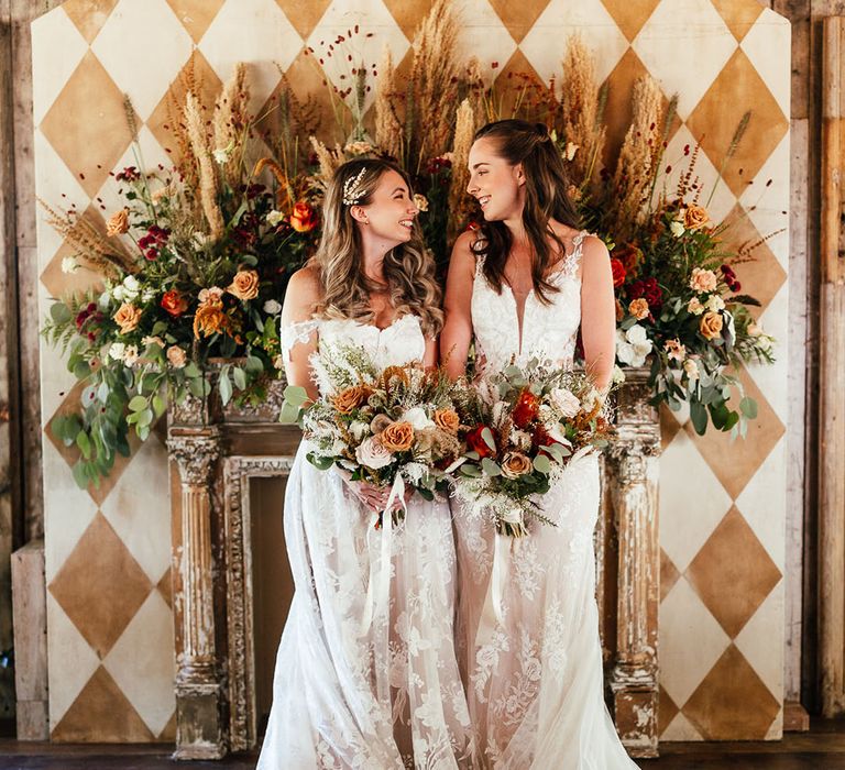 Brides in boho lace wedding dresses with autumnal flower bouquets and an orange rose and dried pampas grass flower arrangement 