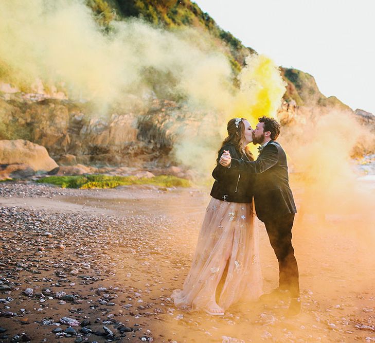 Bride wearing personalised leather jacket kisses groom whilst stood within orange and yellow smoke bombs along the beach