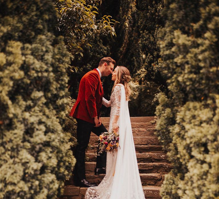 Groom in an orange velvet suit jacket with bride wearing a long sheer cape 