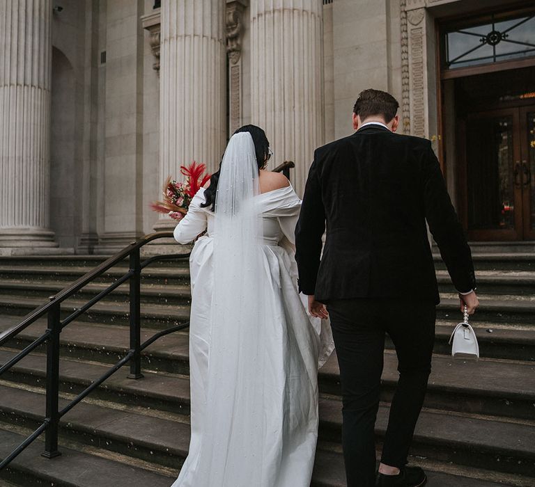 Bride holds bright floral bouquet and wears floor-length veil as she walks with her groom up the stairs of the Old Marylebone Town Hall