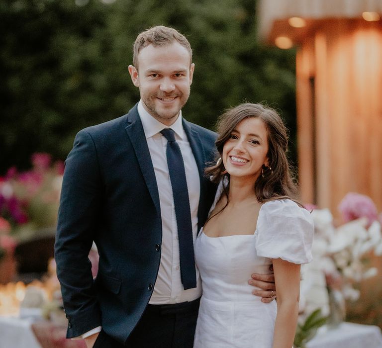 Bride wears white summer dress with puffed sleeves and stands with her groom in blue suit and tie for back garden wedding