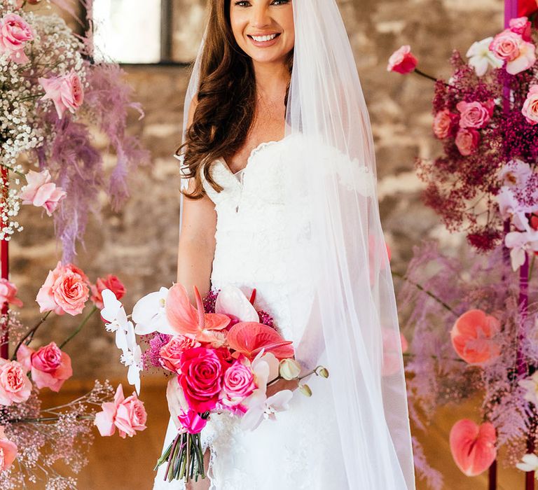 Bride stands between bright pink floral arch and wears lace sweetheart neckline wedding dress with floor-length cathedral veil 