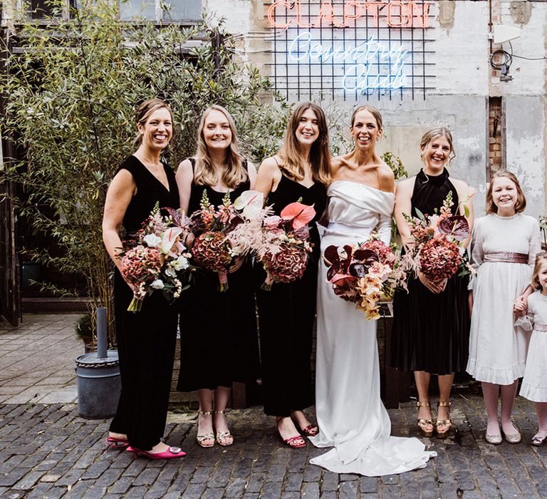 Bridesmaids in black dresses and jumpsuits with flower girls in white and pink ribbon dresses standing next to the bride in a cowl back wedding dress