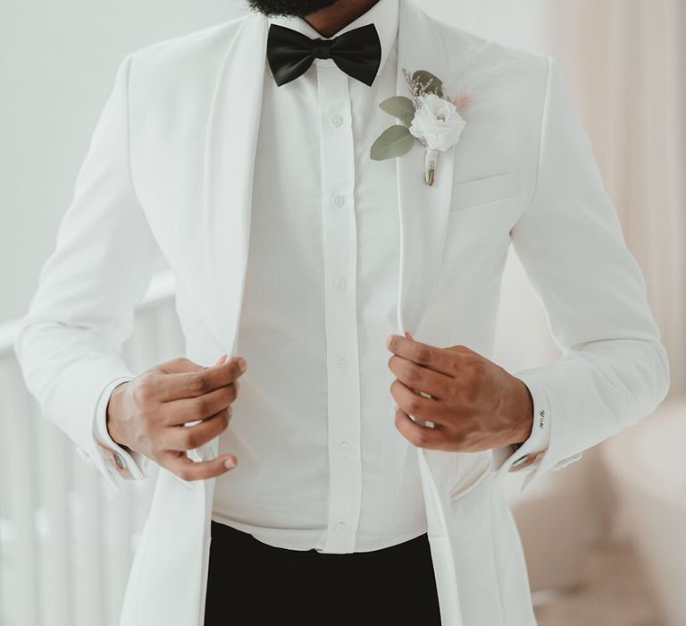 Groom wears white suit jacket with black tie and white floral buttonhole for outdoor wedding ceremony 