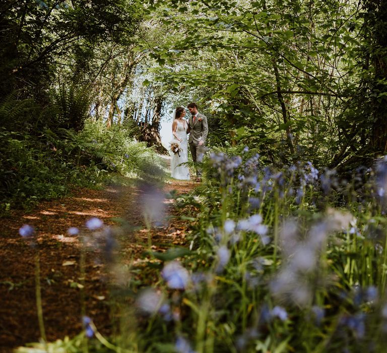 Bride and groom standing amongst the trees at The Green Cornwall wedding venue with light purple flowers in shot