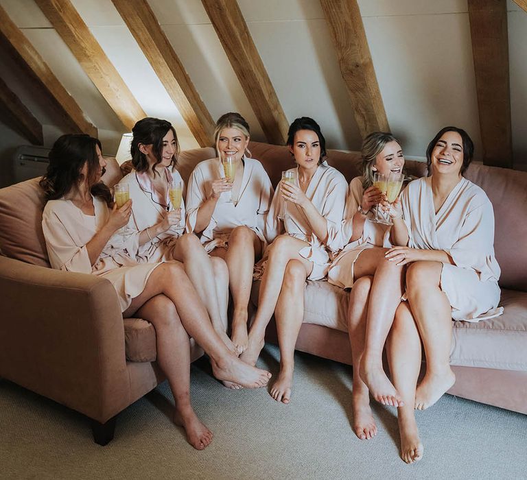 Bridesmaids sit together in their matching pink robes drinking from personalised glasses 