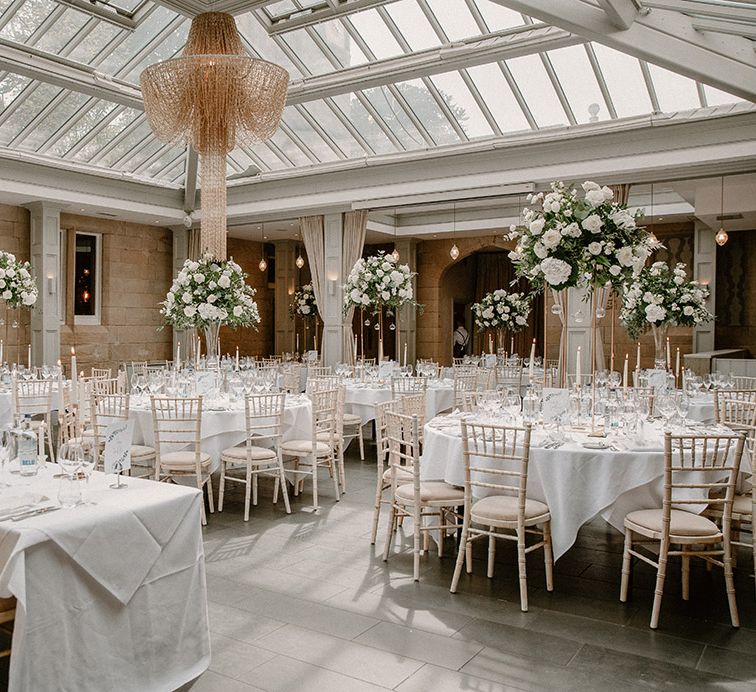 White & green flower installation centrepieces on round tables with white tablecloths as chandelier hangs above at Hampton Manor reception