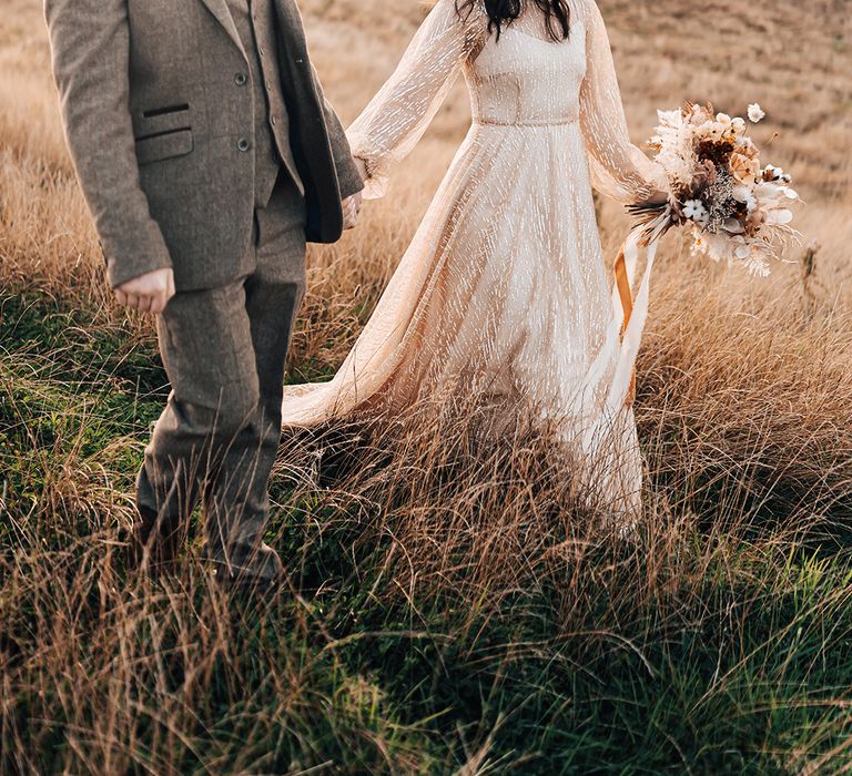 Bride in gold wedding dress walks with her groom in golden fields outdoors 