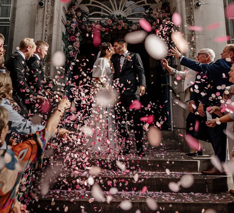 Bride in v neck wedding dress with puff sleeves and front slit and groom in black tux kissing on the steps of Somerset House amidst pink confetti