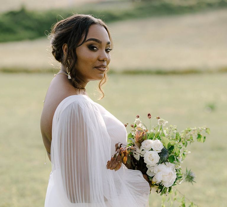 Bride stands in field showing off her backless wedding dress and bridal up do while holding bouquet of roses, thistles and wildflowers