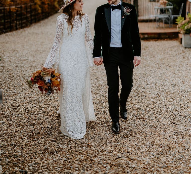 Bride in a boho laser cut lace wedding dress with bell sleeves walking with the groom in a velvet tuxedo and colourful bouquet 