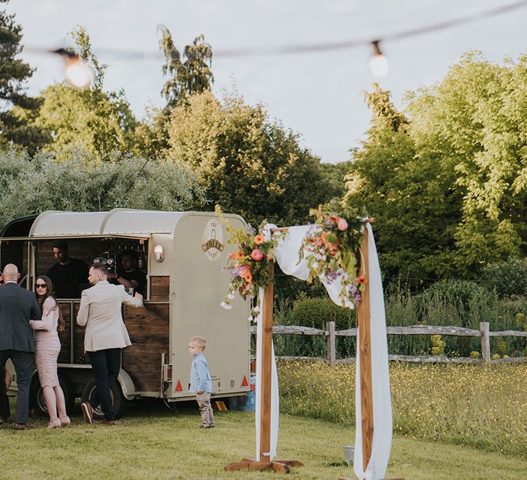 Outdoor truck bar beside wooden arch complete with white drapes and floral decor