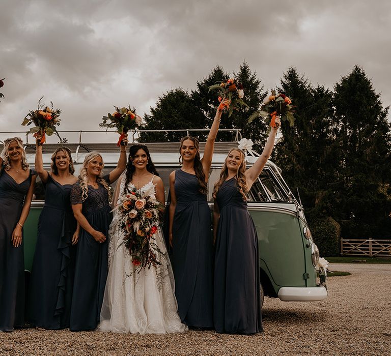 Bridesmaids in navy blue dresses in different styles stand with their bouquets in the air in front of the VW camper van with the bride holding a cascade wedding bouquet