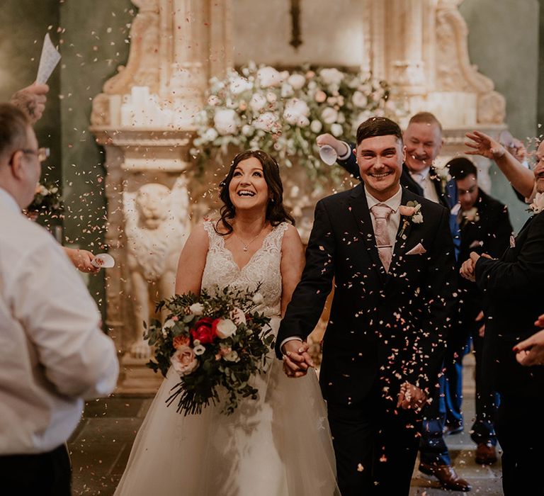 Bride in lace wedding dress walks with her groom through colourful confetti exit 