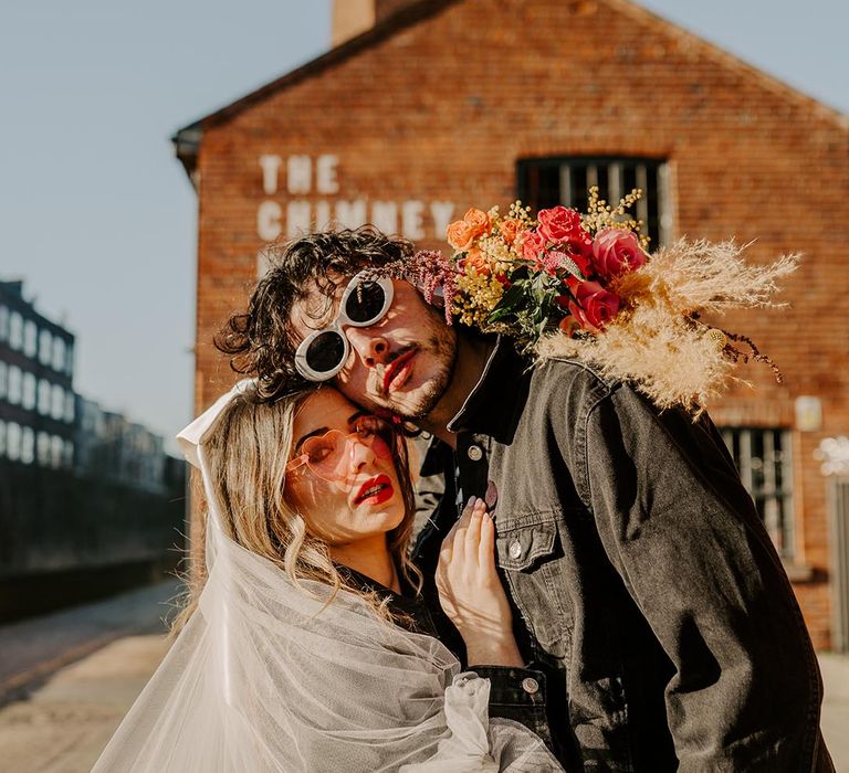 Bride in ruched sheer wedding veil, black leather jacket and pink sheer sunglasses posing with groom in black denim jacket and white sunglasses 