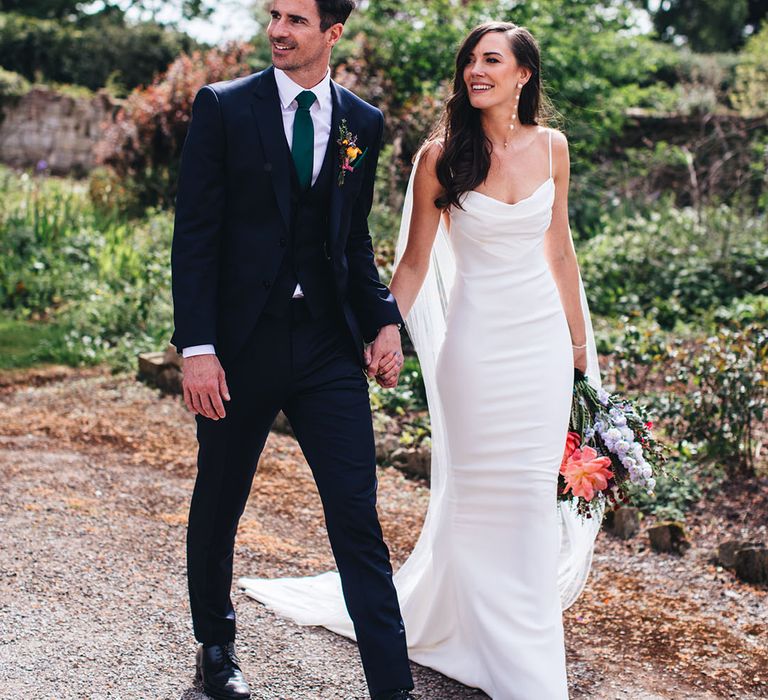 Groom in three piece suit with green tie and bright buttonhole walking with the bride in a fitted satin cowl neck Kyha wedding dress