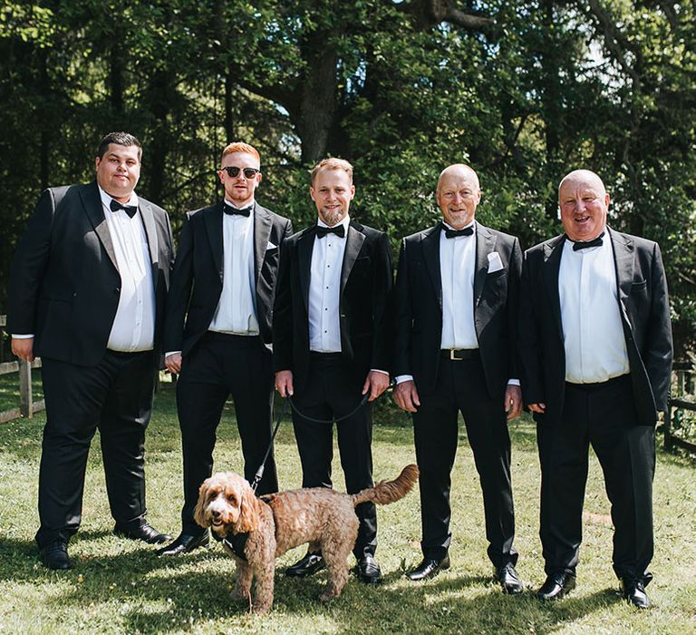 Groom and groomsmen in black tie tuxedos with the pet dog for their group wedding photos 