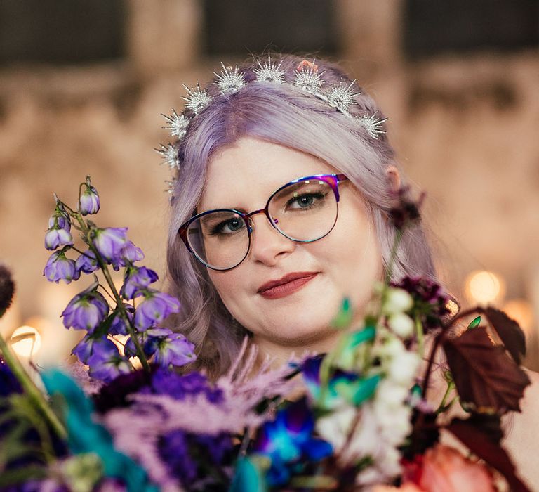 Bride in purple rimmed glasses and celestial silver sparkly bridal headband holding colourful peacock inspired bouquet 