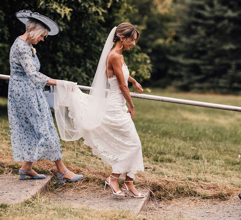 Mother of the bride in blue floral dress with blue hat and shoes with the bride in a fitted lace wedding dress and father of the bride in a black tux walking down stairs on the way to the ceremony