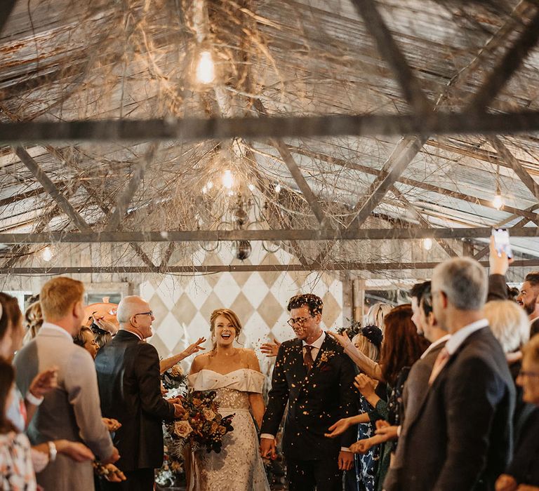 Bride in Love Spell wedding dress walks alongside her groom through colourful confetti exit 