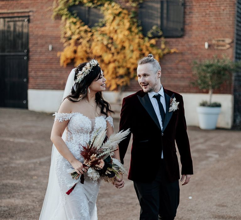 Milling Barn wedding with the bride in an off the shoulder lace wedding dress walking with the groom in a burgundy velvet wedding suit with dried flower bouquet, buttonhole and flower crown