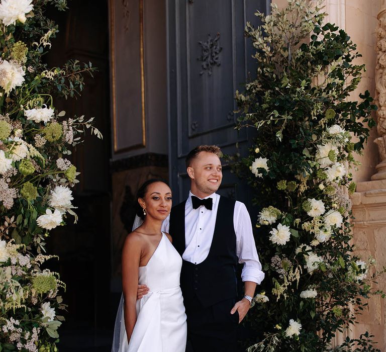 Bride in a one shoulder Jane Hill Bridal gown with front slit and groom in bow-tie at church entrance with column flowers