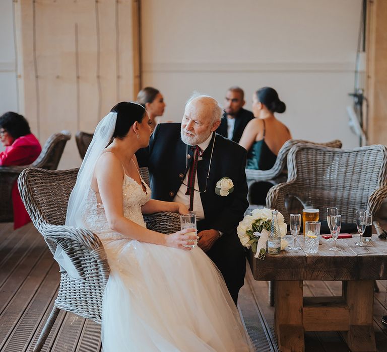 Bride sitting on a wicker chair at Grittleton House wedding venue in lace sleeveless v neck Enzoani wedding dress with tulle finish and church length veil with 3d appliqué embroidered flowers chatting with wedding guest 