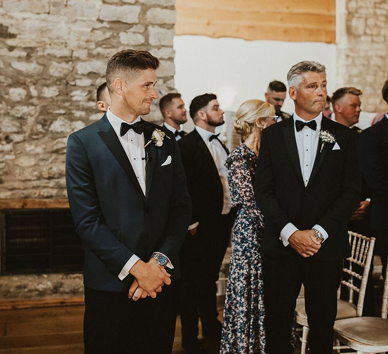 The groom in a traditional tuxedo smiles as he sees the bride for the first time 