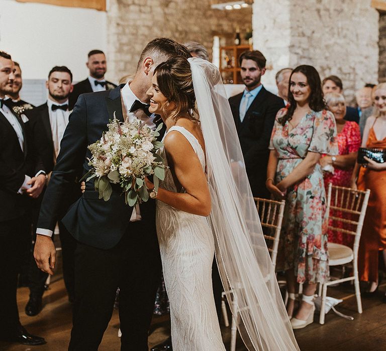 Groom in black tie kisses the bride on the cheek as she greets him after walking down the aisle 