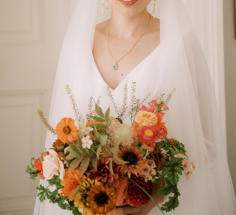 Bride wearing dainty gold jewellery in lace wedding veil and traditional wedding gown holding colourful bridal bouquet 